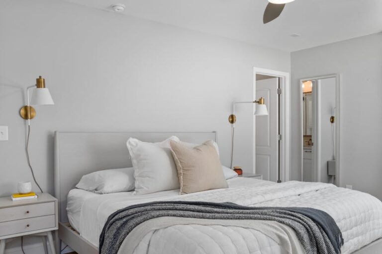 Modern bedroom featuring a neatly made bed with a gray headboard and soft white bedding. There are white pillows, including one beige accent pillow. The room has wall-mounted brass sconces, white nightstands with a lamp, and an open door revealing a bathroom.