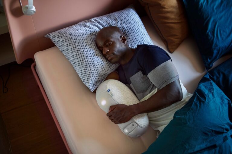 A man lies in bed, sleeping on his side while holding a white, pill-shaped device. He rests his head on a patterned pillow and is covered with a blanket. The setting appears calming and comfortable.