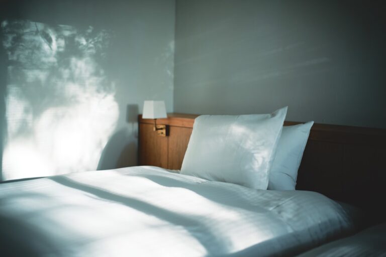 A neatly made bed with white linens is positioned against a wall with soft, natural light filtering in. A wooden headboard features a small, modern reading light on the left side. The overall atmosphere is calm and serene, with subtle shadows cast on the wall.