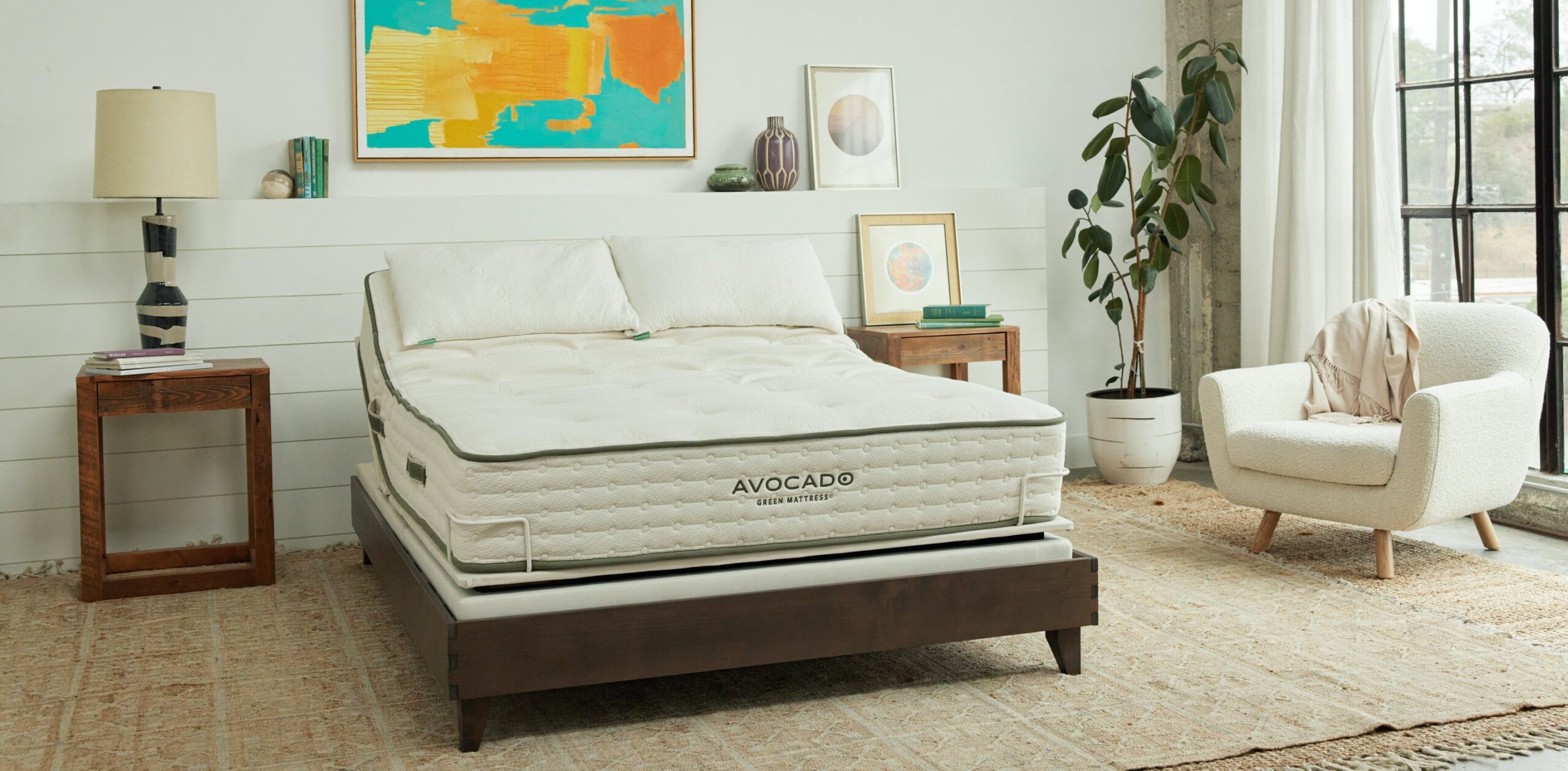 A modern bedroom featuring a neatly made bed with an Avocado mattress, fluffy white pillows, and wooden side tables. The room is decorated with a potted plant, an armchair, and colorful abstract wall art. Natural light streams through large windows with black frames.