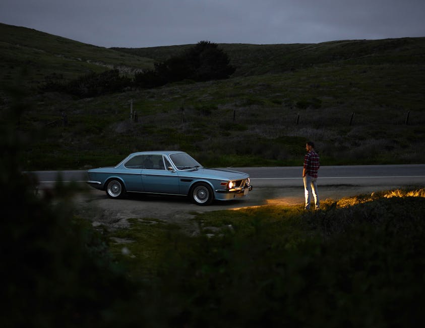 A person stands beside a vintage blue car parked on the side of a desolate road, surrounded by rolling green hills under a dim, overcast sky. The car's headlights illuminate the ground in front, adding a soft glow to the scene.