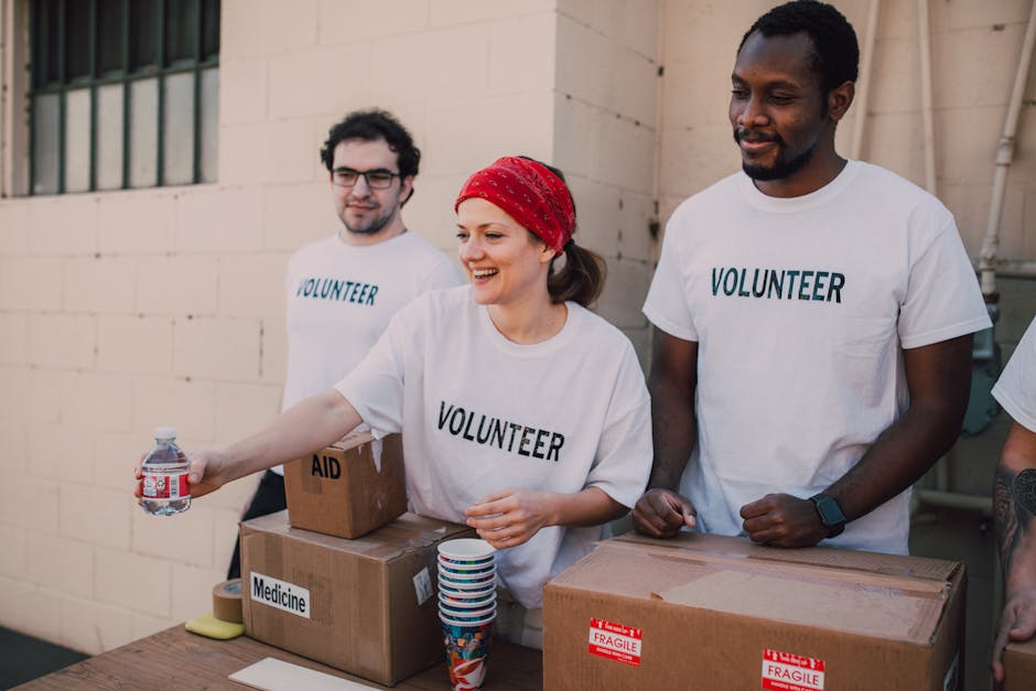 Three volunteers wearing white "Volunteer" t-shirts stand behind a table with donated items, including medicine and water, during a charity event. The person in the middle, wearing a red headband, is handing a bottled water to someone off-camera.