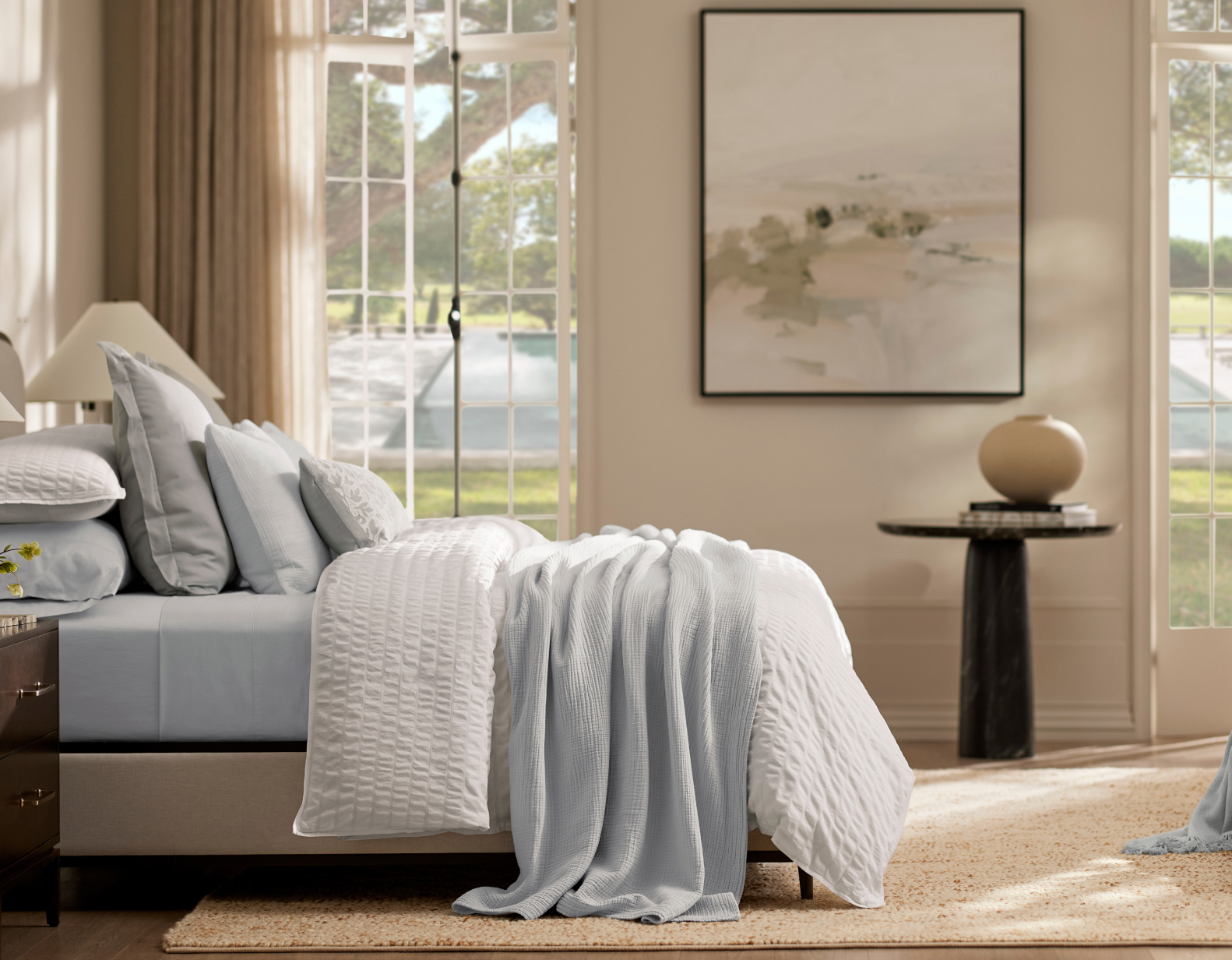 A serene bedroom features a neatly made bed with light blue and white bedding. There's a large rectangular window with a scenic outdoor view, a bedside table with a lamp, an abstract painting on the wall, and a round side table holding a spherical ornament.