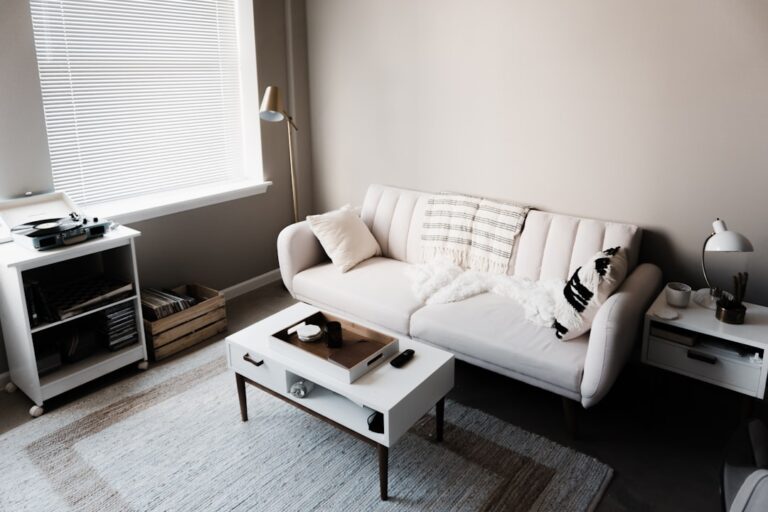 A cozy, neutral-toned living room features a white sofa with pillows and a throw blanket. A small rectangular coffee table sits in front of the sofa, and a floor lamp is positioned in the corner. There's natural light coming in through a window with blinds.