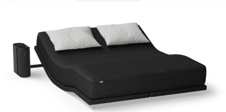 A black adjustable bed with a modern design, featuring two white pillows and a black mattress. The head section is slightly elevated, the foot section curves downward, and there is a small black rectangular device next to the bed, possibly a remote control or speaker.