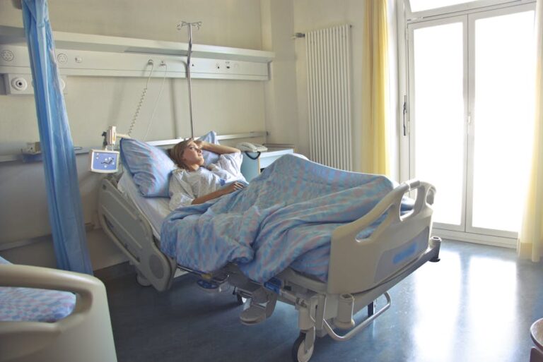 A person lies in a hospital bed covered with blue-patterned sheets. They hold their head with one hand, appearing to be in discomfort. The room has light walls, a window with sheer curtains, and medical equipment by the bed.