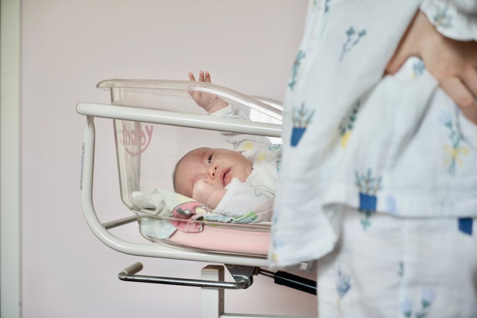 A baby in a hospital bassinet stretches out an arm. The infant is lying on a pink blanket inside a clear plastic crib, with a person in patterned hospital attire standing nearby. The scene suggests a recently born child in a hospital setting.