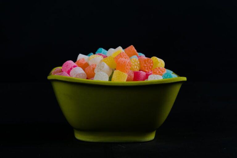 A green bowl filled with a colorful assortment of candy. The candies are various shapes and colors, including pink, yellow, orange, white, and blue. The background is black, highlighting the vivid colors of the candies.