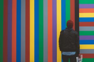 A person wearing a red beanie and a black jacket stands in front of a colorful, vertically striped artwork. The stripes consist of various colors, including red, blue, green, yellow, and gray. The person holds a folded newspaper in their hand.