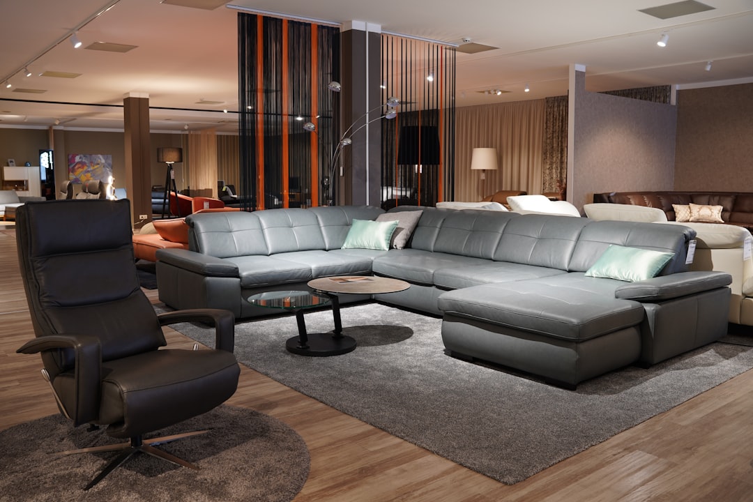 A showroom displaying modern furniture including a large, gray sectional sofa with green pillows, a round black coffee table, and a black recliner chair. The space features wooden floors, warm lighting, and various other furniture pieces in the background.