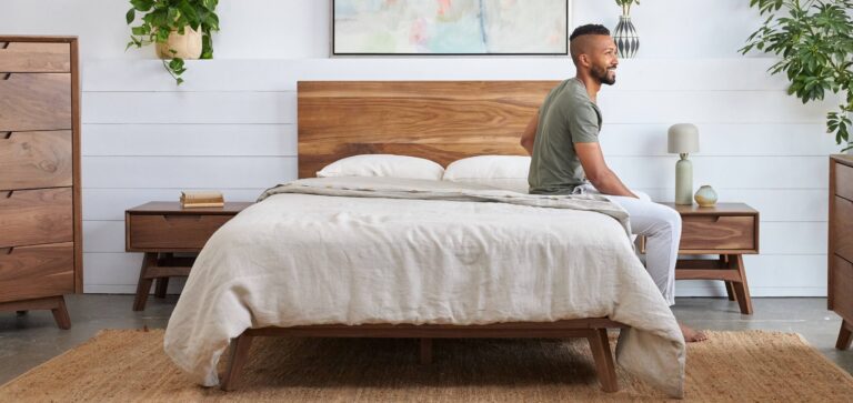 A person sits on the edge of a wooden bed with beige bedding in a modern, minimalist bedroom. The room includes wooden furniture, a rug, indoor plants, and a painting above the bed. The decor is simple with a neutral color palette.