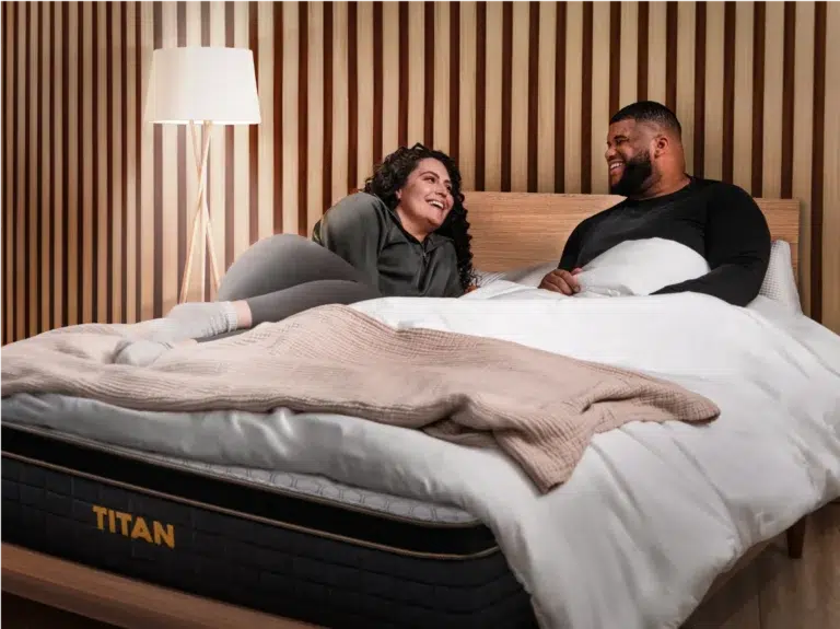 A woman and a man are lying on a bed in a cozy room with wooden paneling on the walls and a lamp in the background. They are smiling and looking at each other. The bed, labeled "Titan Plus Elite," is neatly made with white bedding, and a blanket is draped over the foot.