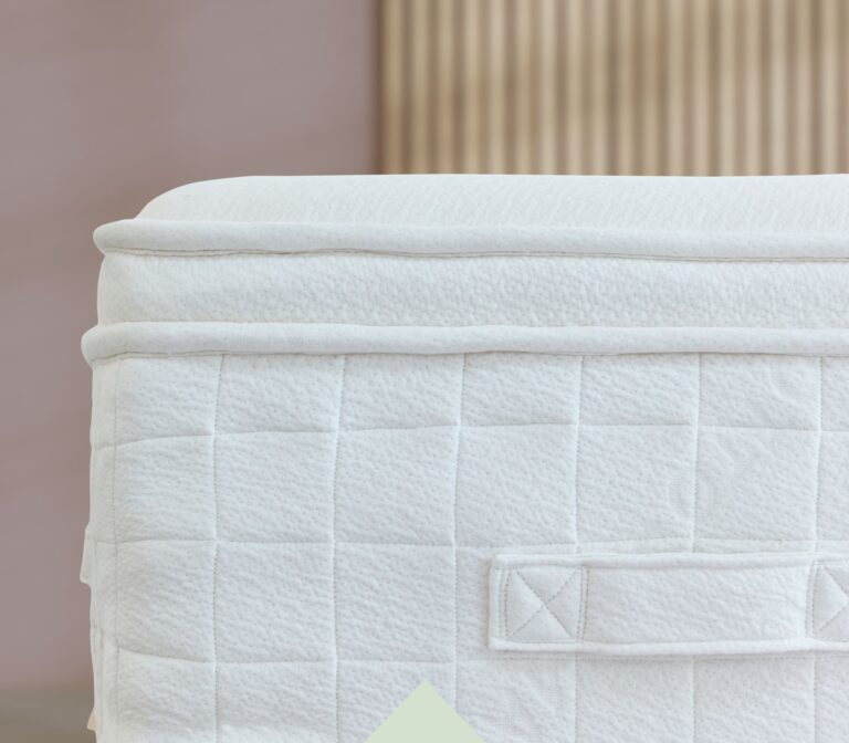 Close-up of a white mattress's side, featuring a quilted pattern with vertical and horizontal stitched lines forming square shapes. A fabric handle is visible on the side, designed for carrying or adjusting the mattress. Background is blurred with beige tones.