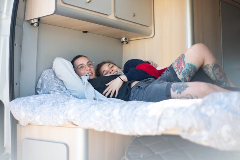 Two people are lying on a bed inside a camper van, smiling and cuddling. The person on the left is wearing glasses and a white sweater, and the one on the right has tattoos on their legs and is wearing a black and red sweater along with gray shorts.