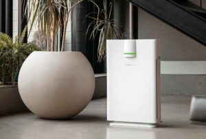 A modern, sleek white air purifier with a green power indicator light stands on a polished concrete floor next to a large, round, white planter containing tall plants. The scene is set in a contemporary indoor space with industrial elements.