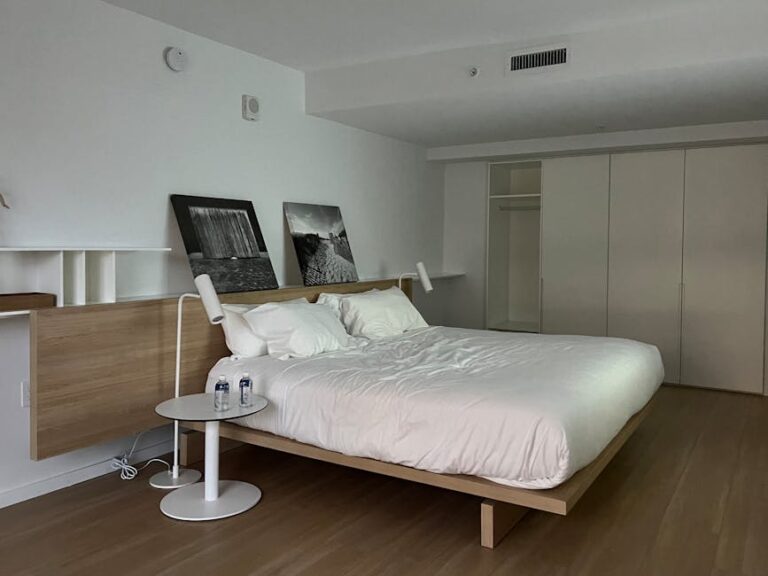 A minimalist bedroom with a large wooden bed, white bedding, and two framed black and white photos resting on a ledge above the bed. There are two white table lamps on either side and a small white side table with two water bottles. The room has light wood floors.