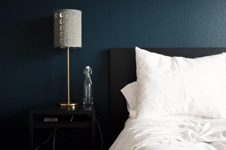 A modern bedroom with a dark, sleek bedside table holding a stylish gold lamp with a perforated shade and a glass water bottle. The bed has crisp white linens and pillows against a deep green wall, creating a serene and elegant atmosphere.