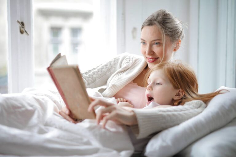 A woman with light-colored hair and a child with red hair are lying in bed by a window, both under a white blanket. The woman is holding a book and reading to the child, who is attentively listening and looking at the pages.