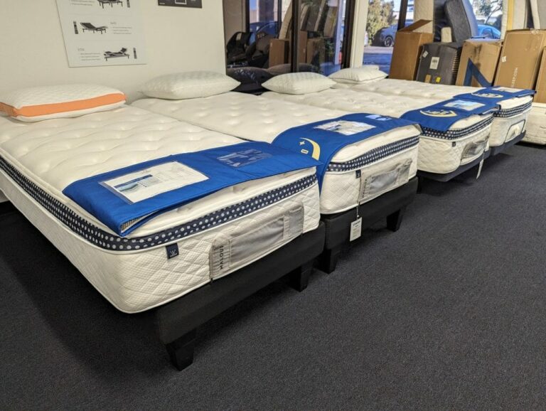 Four mattresses lined up side by side on display in a store. Each mattress has a blue cover and labeled brochure on top. The mattresses are on box springs and bases, with some pillows placed on them. The background shows posters and cardboard boxes.