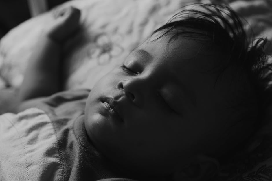 A black-and-white close-up photo of a sleeping baby. The baby lies on a soft surface with eyes closed and slightly parted lips. Their hair is tousled, and their hand rests near their head. The setting appears peaceful and the light is soft, casting gentle shadows.