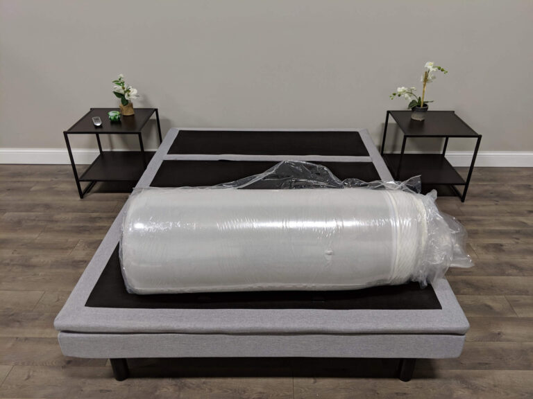 A neatly arranged bedroom with a queen-sized bed frame has a rolled-up mattress in vacuum-sealed packaging placed in the center. The bed is flanked by two black nightstands, each holding a small plant and decorative item. The flooring is wooden.