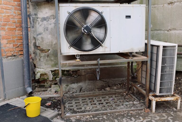 A large outdoor air conditioning unit mounted on a metal frame stands against a weathered brick and concrete wall. To the right, a smaller, weathered unit is partially visible. A yellow bucket sits on the ground nearby. The area is paved with interlocking bricks.