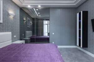 Modern bedroom with gray walls and a purple bedspread. The room features a large mirrored closet, a small nightstand beside the bed, and a wall-mounted TV. Recessed lighting and a patterned wallpaper add a contemporary touch to the space.