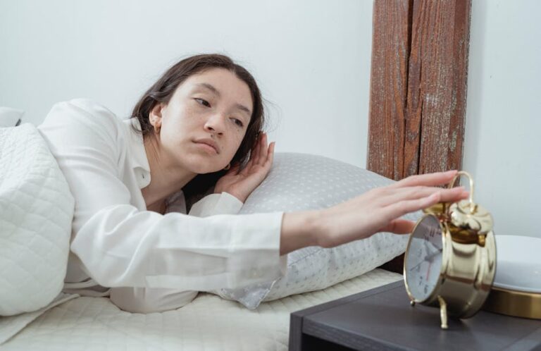 A person lies in bed reaching out to turn off a gold-colored alarm clock on a bedside table. They look tired and are dressed in a white long-sleeve shirt with a white blanket draped over them. The room has a white wall and a wooden beam in the background.