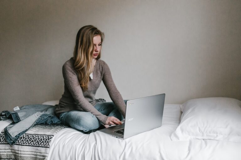 A person with long hair, wearing a brown sweater and jeans, is sitting on a bed and typing on a silver laptop. The bed has white sheets and pillows, with a patterned blanket and a denim jacket lying next to the individual. The room has plain, light-colored walls.