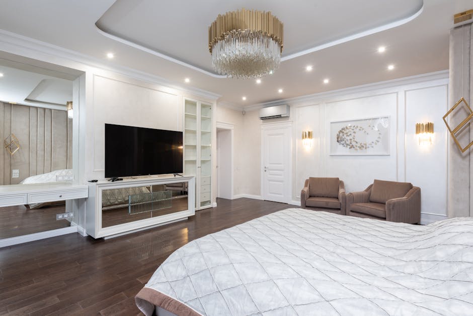 A modern, well-lit bedroom with a large bed covered in a grey quilt. The room features a large flat-screen TV on a console, a chandelier, two brown armchairs, a mirrored wall, and minimalistic decor. The wooden floor adds warmth to the otherwise neutral palette.