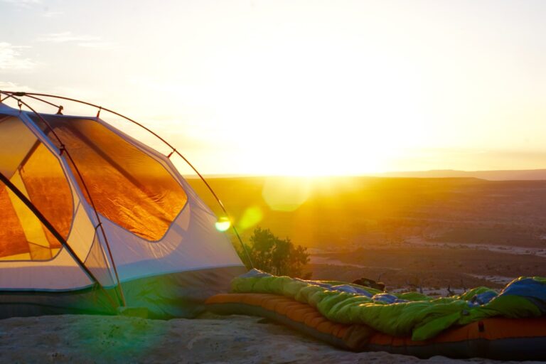 A tent pitched on a sandy landscape with a colorful sleeping bag beside it, basking in the glow of the sunrise. The horizon shows arid terrain and distant plateaus, creating a serene and inviting camping scene in the early morning light.