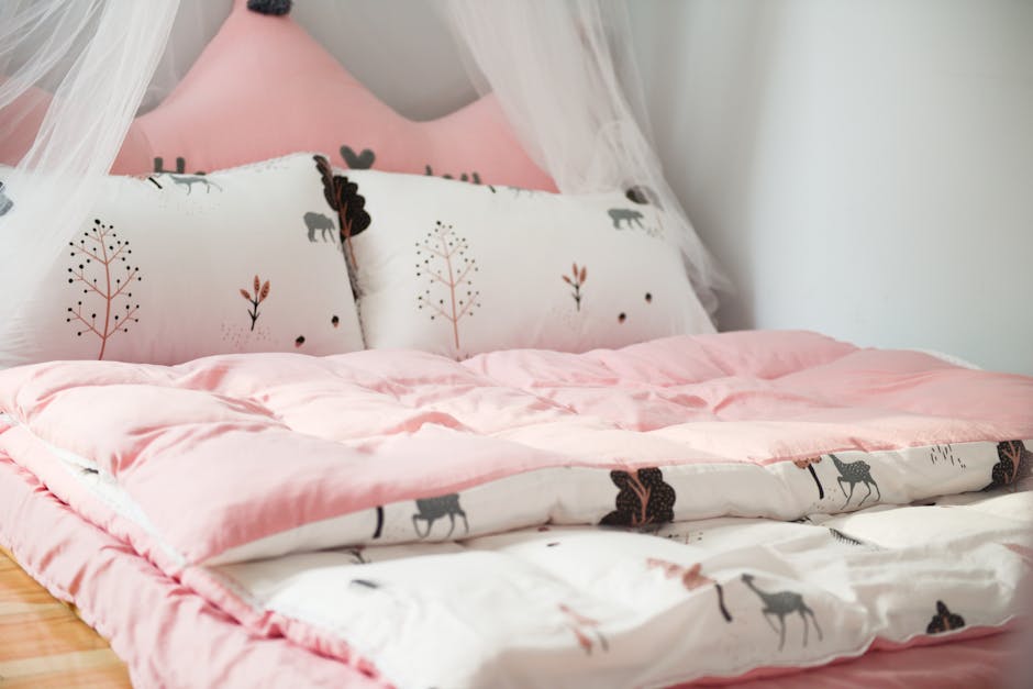 A cozy bed with light pink bedding and a padded pink headboard. The bed is adorned with pillows and a comforter featuring animal and tree motifs. A white mosquito net is partially seen hanging above the bed, adding a dreamy touch to the serene setup.