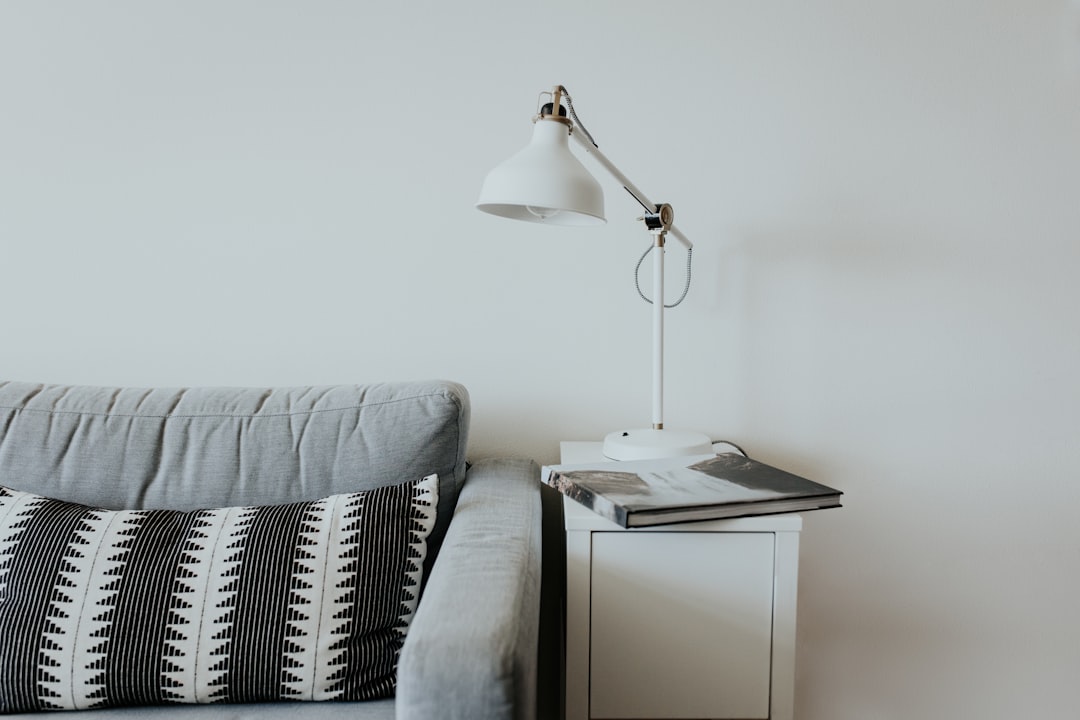 A minimalistic living room setup featuring a light gray sofa with a patterned black-and-white cushion, next to a white side table. On the table is a white desk lamp and a couple of magazines. The background wall is plain and light-colored.