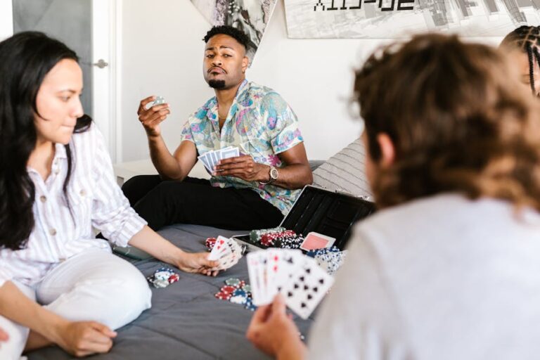 A group of people sit on a gray bedspread, playing a card game. One person in the center is holding cards and appears to be looking thoughtfully. There are poker chips and a case on the bedspread. A bright window is visible in the background.