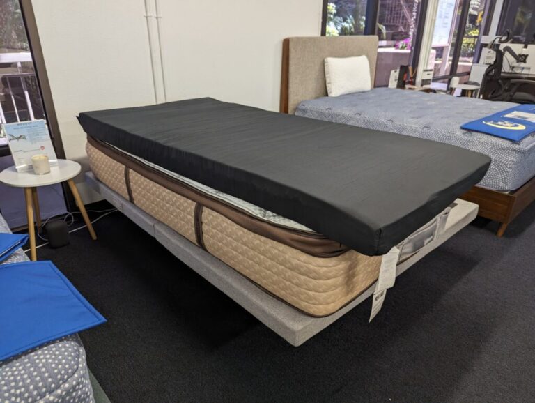 A showroom displays a bed with a brown mattress and a black mattress topper. The bed frame is light gray and adjacent to a small, round white side table with a candle. Another bed with a blue mattress is visible in the background near large windows.