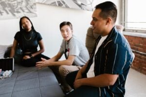 Three people are sitting on a sofa, engaged in conversation. One person (on the left) is wearing a black top, the middle person is wearing a gray shirt, and the person on the right is in a striped shirt. A briefcase is on the table in front of them.