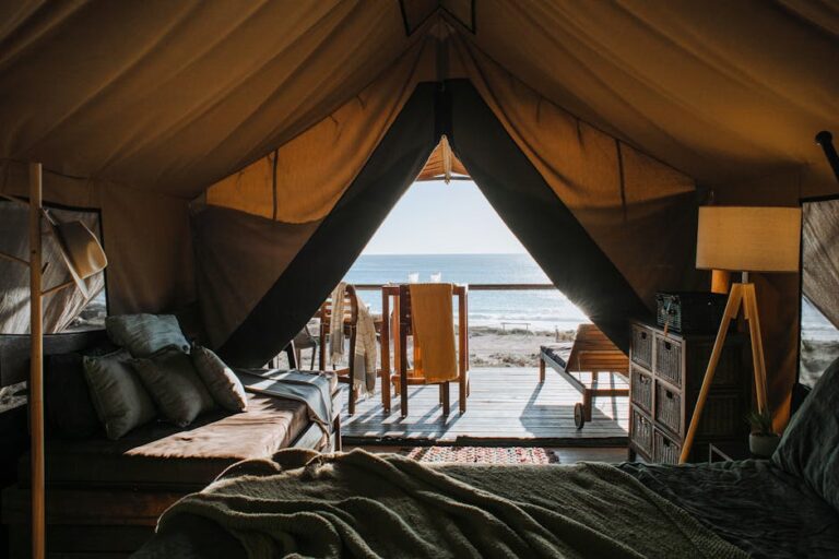 View from inside a cozy glamping tent overlooking a beach. The tent has a bed with pillows, a small bookshelf, and a lamp. The tent opens to a wooden deck with chairs and a table, leading out to the sandy beach and the ocean beyond, under a bright sky.