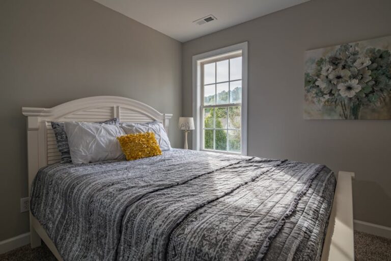 A cozy bedroom with a large window letting in natural light. The room features a neatly made bed with gray and white patterned bedding, white pillows, and a yellow accent pillow. A floral painting hangs on the wall next to the window, adding a touch of warmth.