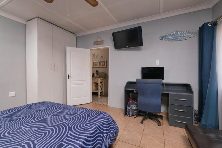 A neatly organized bedroom with a wall-mounted TV, large wardrobe, and a dark-colored desk with a comfortable office chair. The bed has a blue-patterned comforter, and a sign above the door reads "Gents Room." The door is open, providing a view into another room.