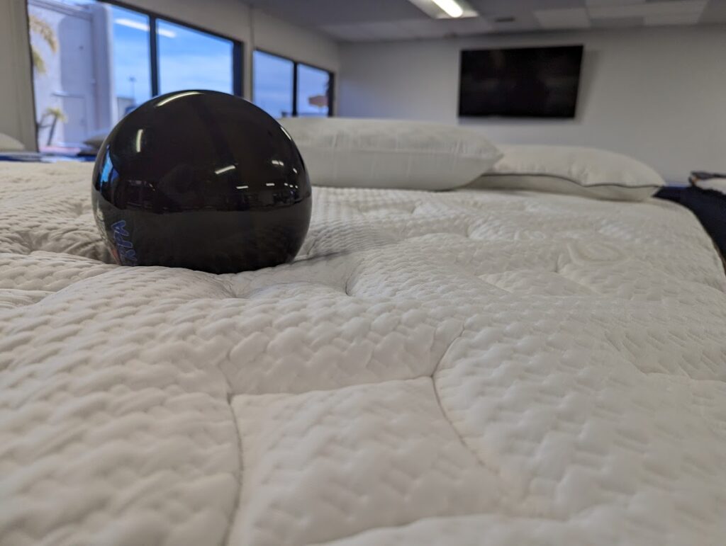 A black spherical object is placed on a white quilted mattress in a brightly lit room with large windows. The foreground of the image focuses on the texture of the mattress, and a television is visible in the background on the wall.