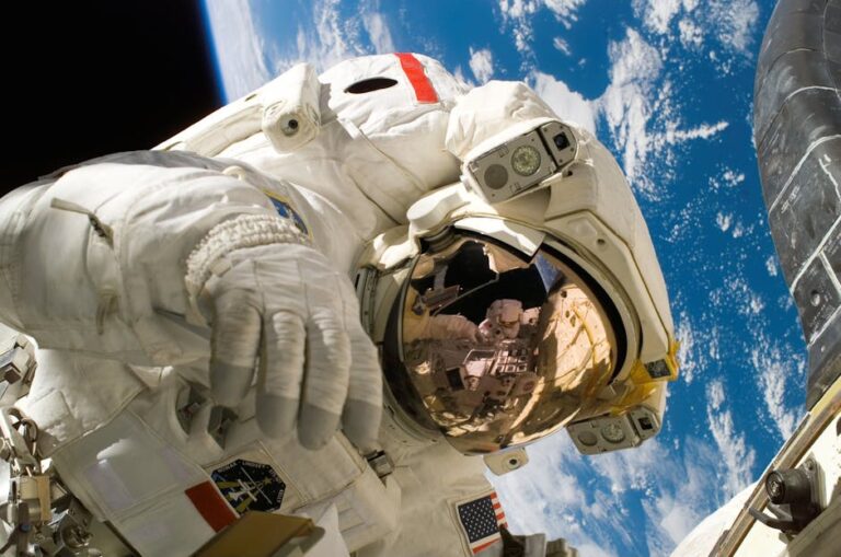 An astronaut in a white spacesuit floats outside a spacecraft with Earth visible in the background. The astronaut's helmet visor reflects part of the spacecraft and another astronaut's gloved hand. The blackness of space frames the Earth below.