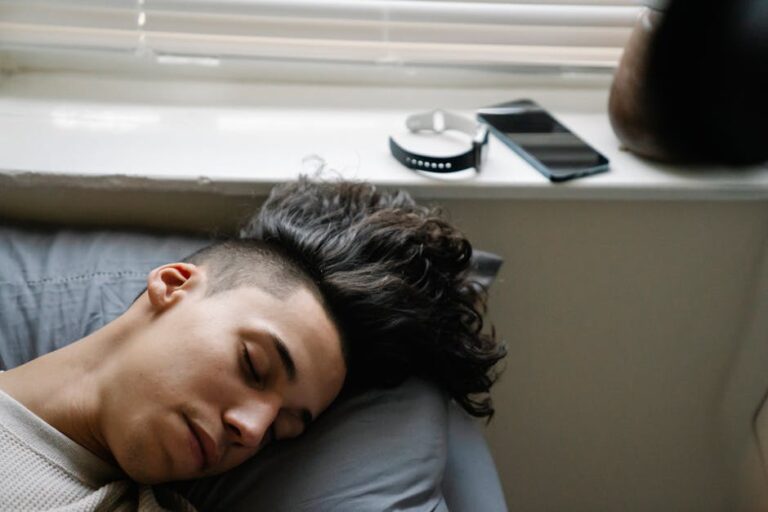 A person with dark, curly hair is sleeping on a pillow near a window with closed blinds. A smartphone and a circular electronic device are placed on the window sill above their head. The person appears peaceful and is wearing a light-colored shirt.