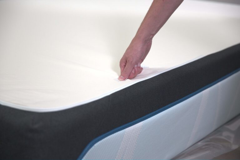 A close-up of a hand adjusting the corner of a white bed sheet on a mattress with a black and blue edge. The sheet is being tucked in neatly to ensure a smooth and fitted appearance.