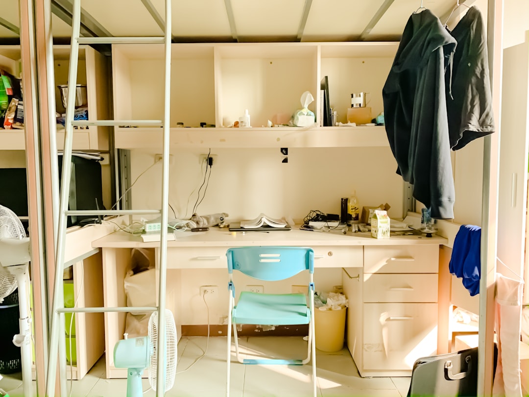 A cluttered study area below a loft bed. The desk is filled with various items, including books, electronics, and a half-empty milk carton. A blue chair is placed in front of the desk, and a black jacket and hoodie are hanging on hooks to the right. A white fan is on the floor.