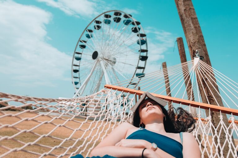 A person relaxes on a hammock with a book covering their face on a sunny day. In the background, a large Ferris wheel and clear blue sky are visible. The scene gives a feeling of leisure and tranquility.