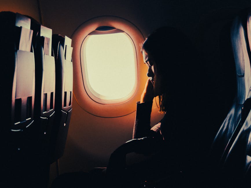 A person sits alone by an airplane window, looking outside with their head resting on their hand. The cabin is dimly lit, creating a contemplative mood. The seats adjacent to the person are empty. The bright sky contrasts with the dark interior.