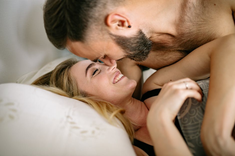 A loving couple in an intimate setting, with a man leaning over a woman who is lying on her back. Both are smiling and appear to be enjoying a tender moment together. The woman is wearing a black bra, and the man has a beard and a pierced ear.