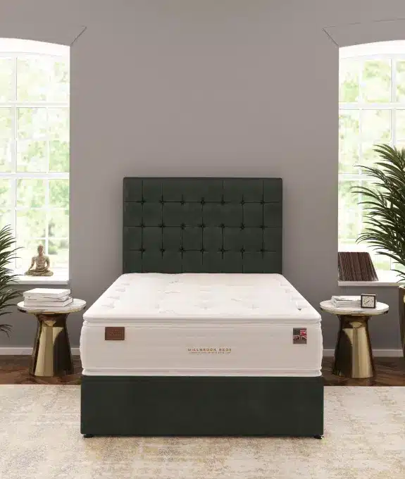 A bedroom features a large bed with a dark green upholstered headboard and a white mattress. On either side, there are small round side tables with various decor items, and large windows behind the bed let in natural light. The walls are gray, and there are plants in the room.