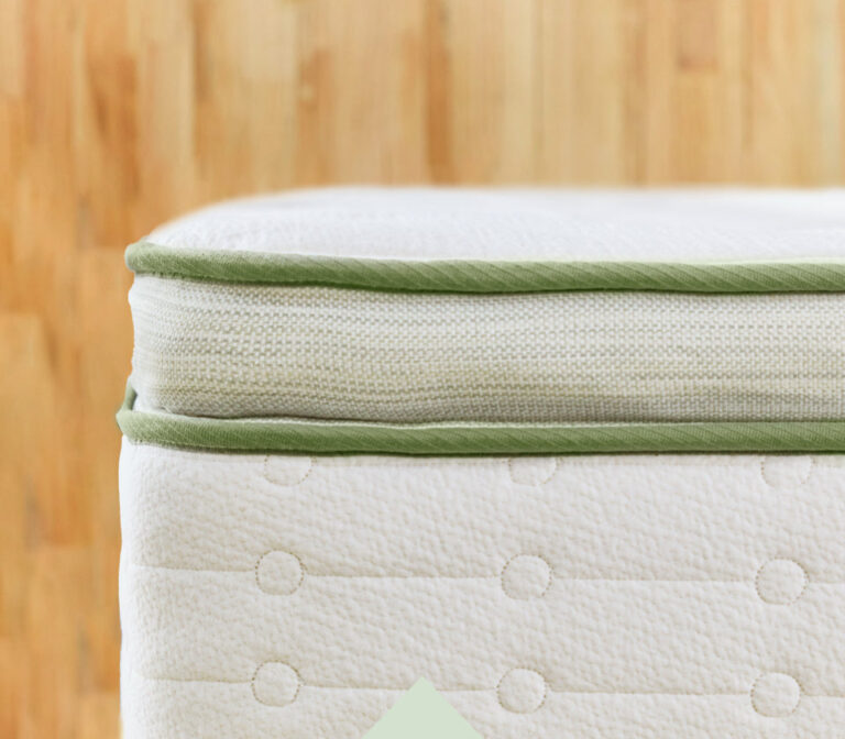 Close-up of the corner of a mattress with a white fabric exterior and light green piping. The background features a wooden floor, adding a warm contrast to the soft-colored mattress. The mattress surface shows a subtle pattern of circles.