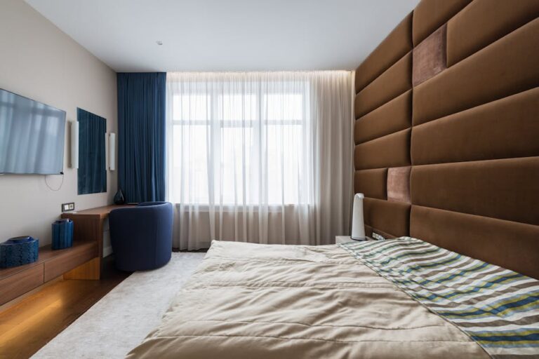 A modern bedroom features a beige bed with striped bedding, a large cushioned brown headboard wall, and a wooden desk with a blue chair. A flat-screen TV is mounted on the wall, and large curtained windows allow natural light to fill the room.